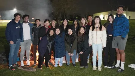 Group of sixteen students smiling in front of campfire outside