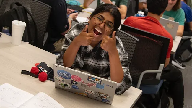 Smiling female student holding two thumbs up sitting at desk with laptop